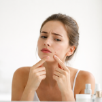 donna che tocca un brufolo sul viso davanti allo specchio mentre controlla la pelle con prodotti skincare sullo sfondo