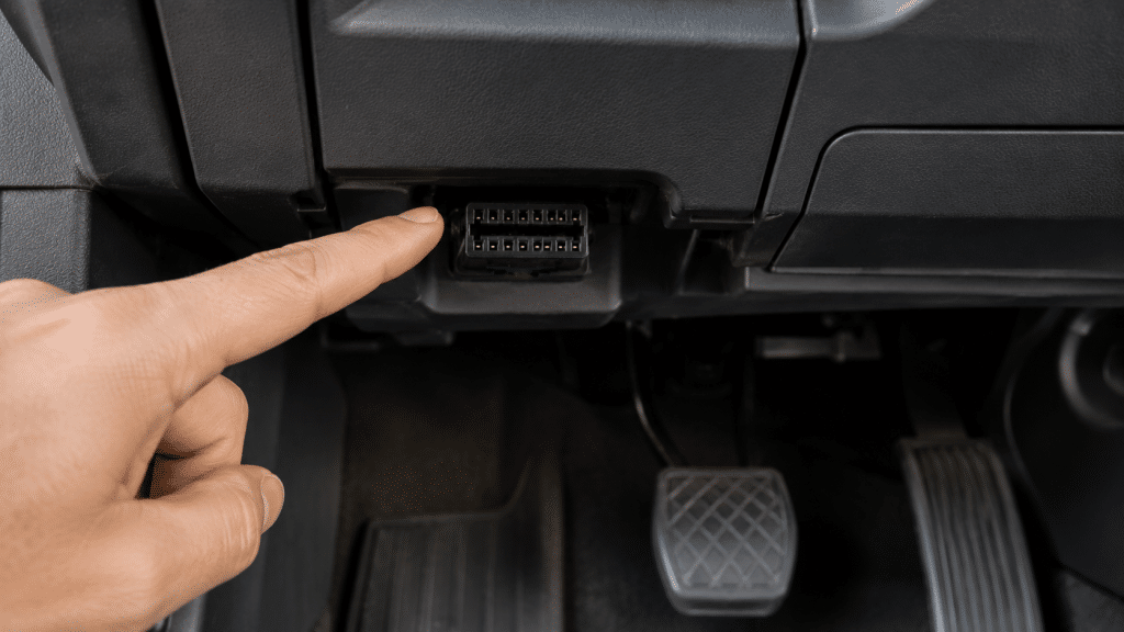 Close-up of the OBD2 socket under the steering wheel of a modern car with a hand pointing to the diagnostic connector.